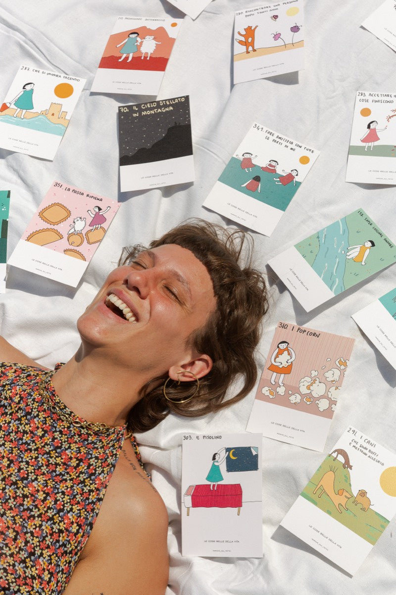 Une personne rit les yeux fermés, allongée sur un drap blanc parmi de nombreuses cartes postales représentant Les Belles Choses de la Vie.